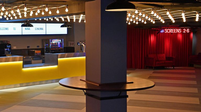 Modern cinema interior with a vibrant yellow bar LED grid light ceiling featue and a red seating area in the background.